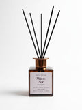 Maison Noir Reed Diffuser with a square amber glass bottle, a metallic rose gold collar, and six black reed sticks, set against a plain white background.
